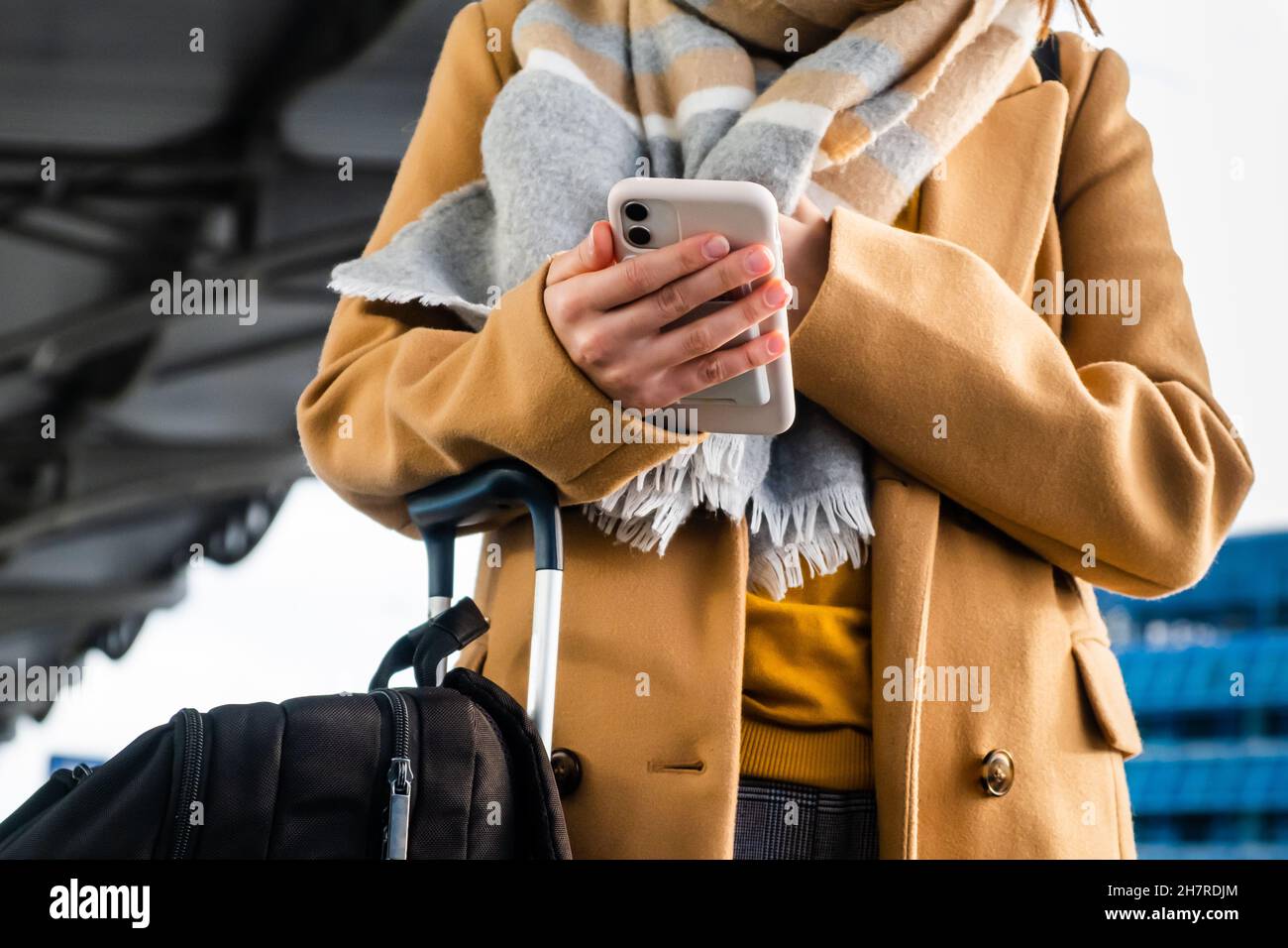Il passeggero Lady in elegante cappotto con sciarpa di lana controlla l'orario del volo sul telefono tenendo la valigia nel corridoio del terminal dell'aeroporto internazionale in primo piano Foto Stock