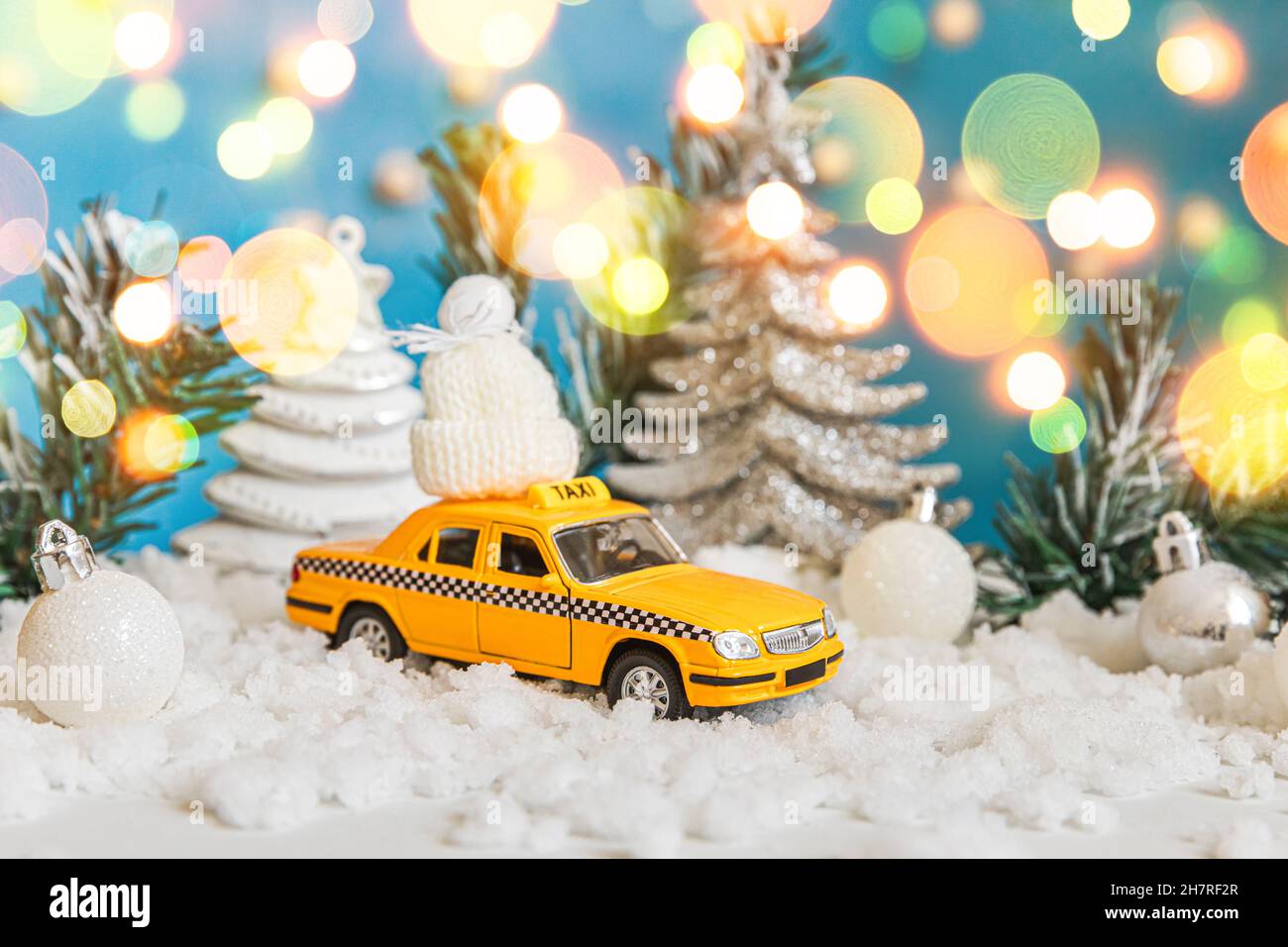 Sfondo banner di Natale. Auto giocattolo gialla Taxi Cab modello e decorazioni invernali ornamenti su sfondo blu con neve e luci di ghirlanda defocused. Concetto di servizio taxi di consegna del traffico cittadino Foto Stock