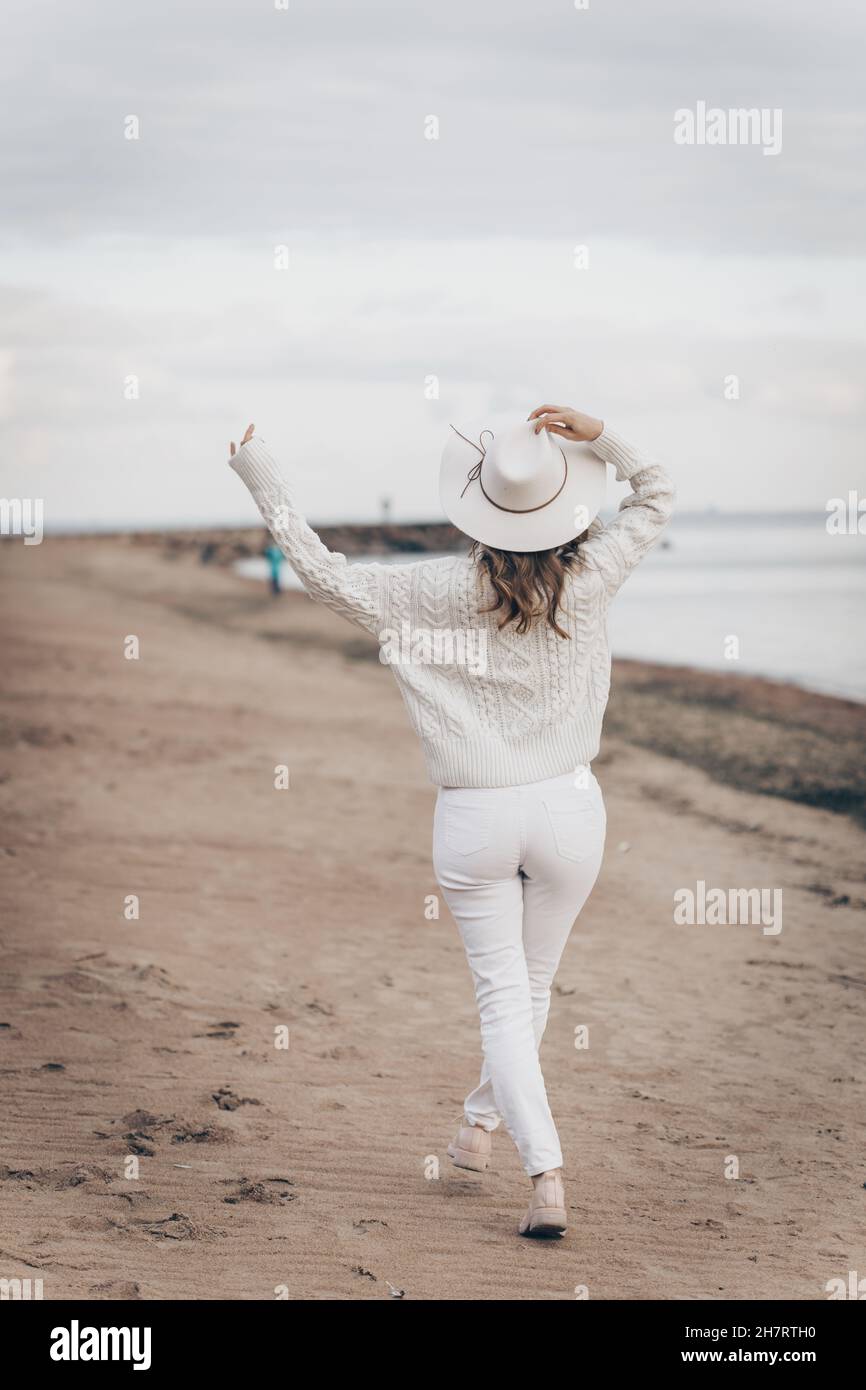 una ragazza bionda in un cappello bianco cammina lungo la riva del mare in un giorno d'autunno Foto Stock