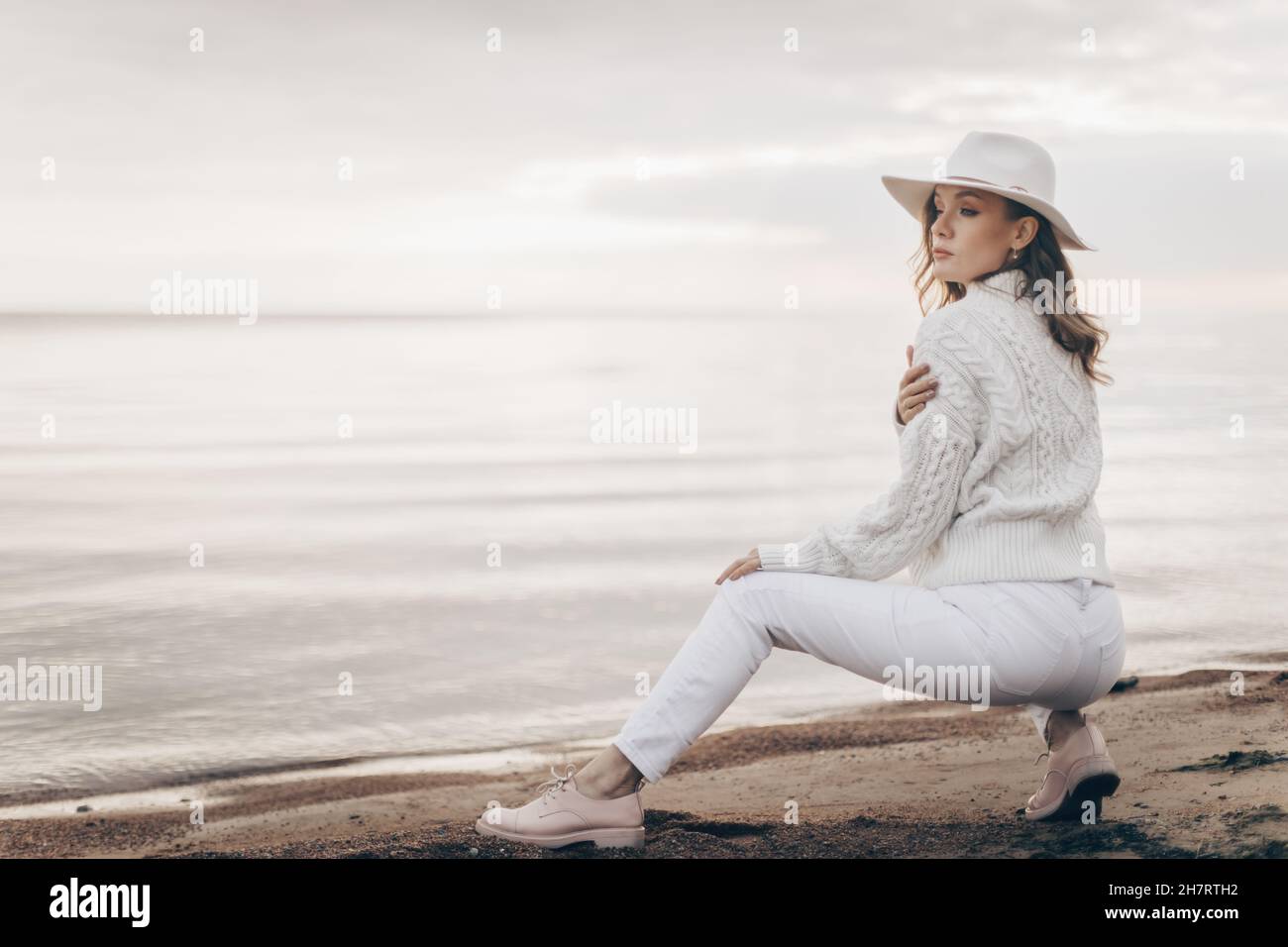 una ragazza bionda in un cappello bianco cammina lungo la riva del mare in un giorno d'autunno Foto Stock
