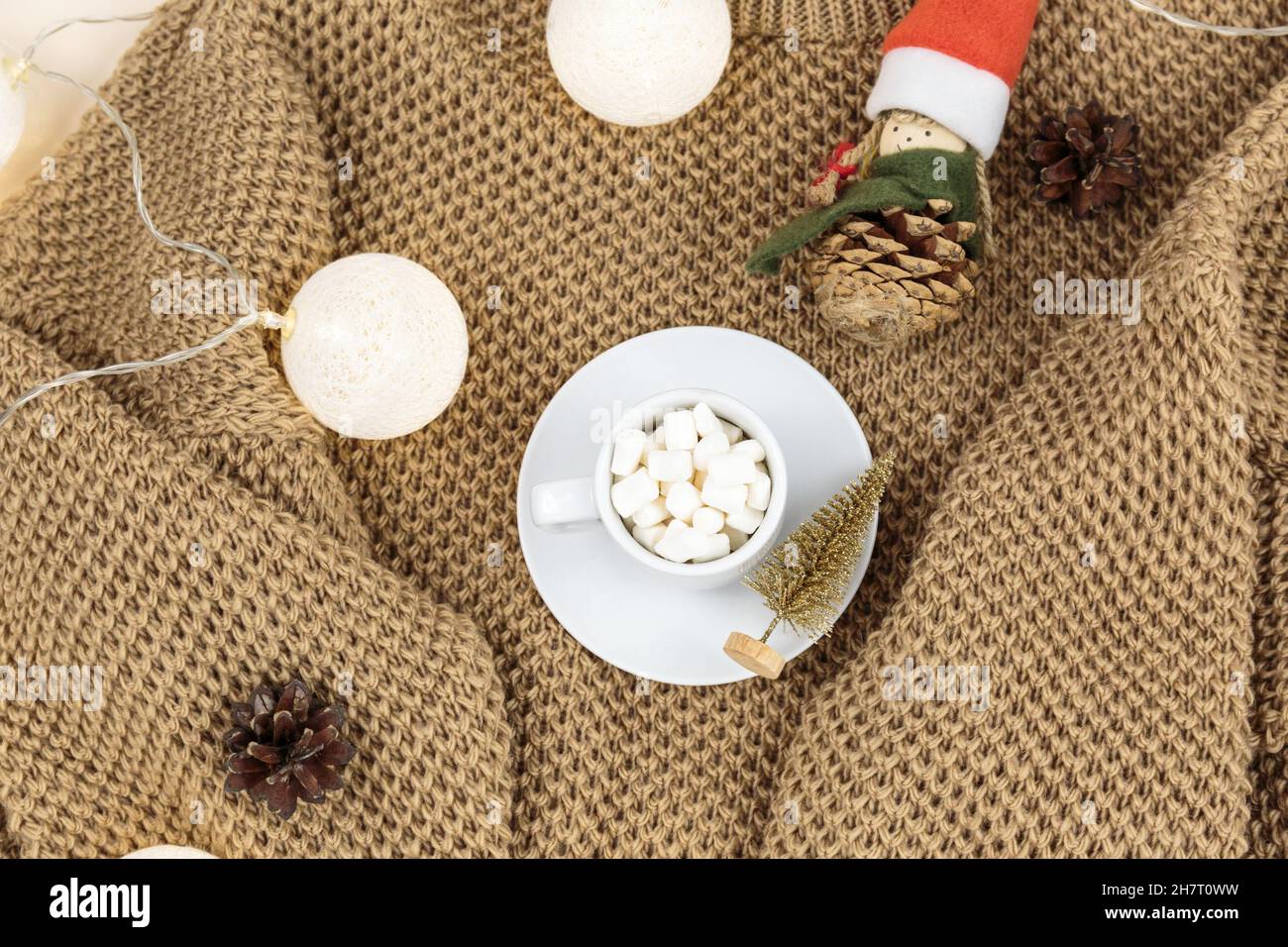 Tazza di caffè, scatola regalo e maglioni in maglia, luci a LED su sfondo beige. Stile Higge, concetto invernale. Accogliente scrivania. Capodanno e Natale Foto Stock