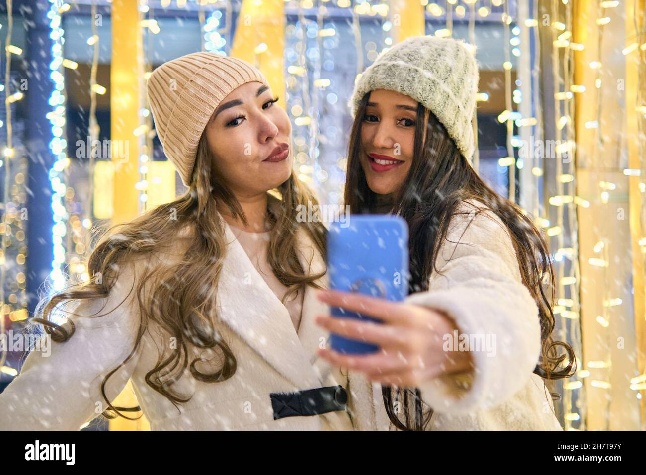 Due ragazze asiatiche ragazze in cappotti bianchi stanno avendo divertimento hugging e selfie sul telefono cellulare, nuovo anno, lampadine Foto Stock