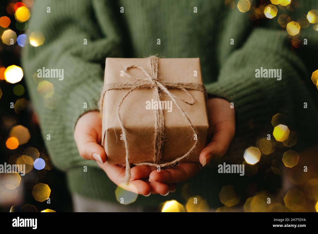 Le mani femminili che tengono la scatola del regalo piacevole, la luce blured del bokeh sullo sfondo. Donna che dà avvolto in carta nera presente. Concetto di zero rifiuti Foto Stock