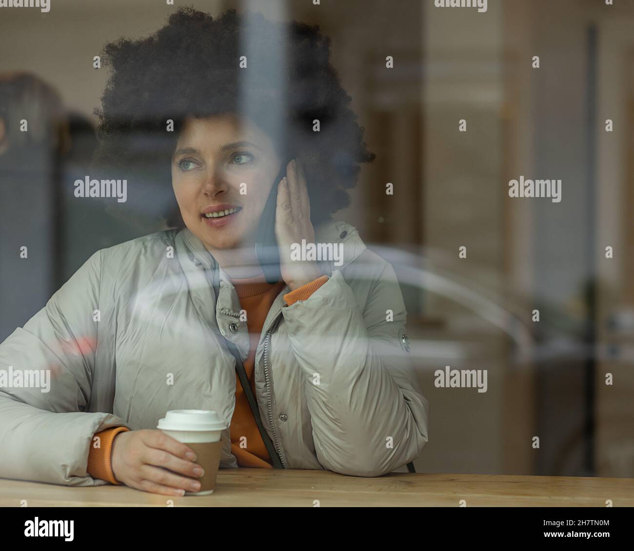 Afro hairstyle donna bianca 40 anni che parla con smartphone al caffè con una tazza di caffè, fotografato attraverso Window. Foto Stock