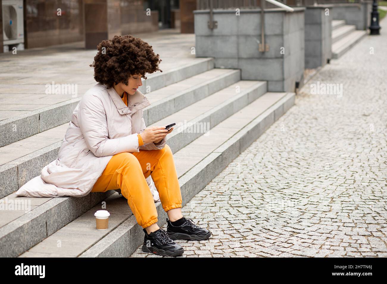Ritratto di giovane, carina donna caucasica 40 anni, Sport abiti casual, seduta su gradini, tazza di caffè monouso. Acconciatura afro. Massaggio texting Foto Stock
