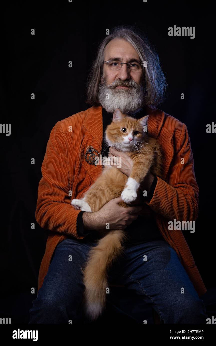 Intellettuale bearded grigio-pelato di età matura che si posa con un gatto adattato dello zenzero Foto Stock