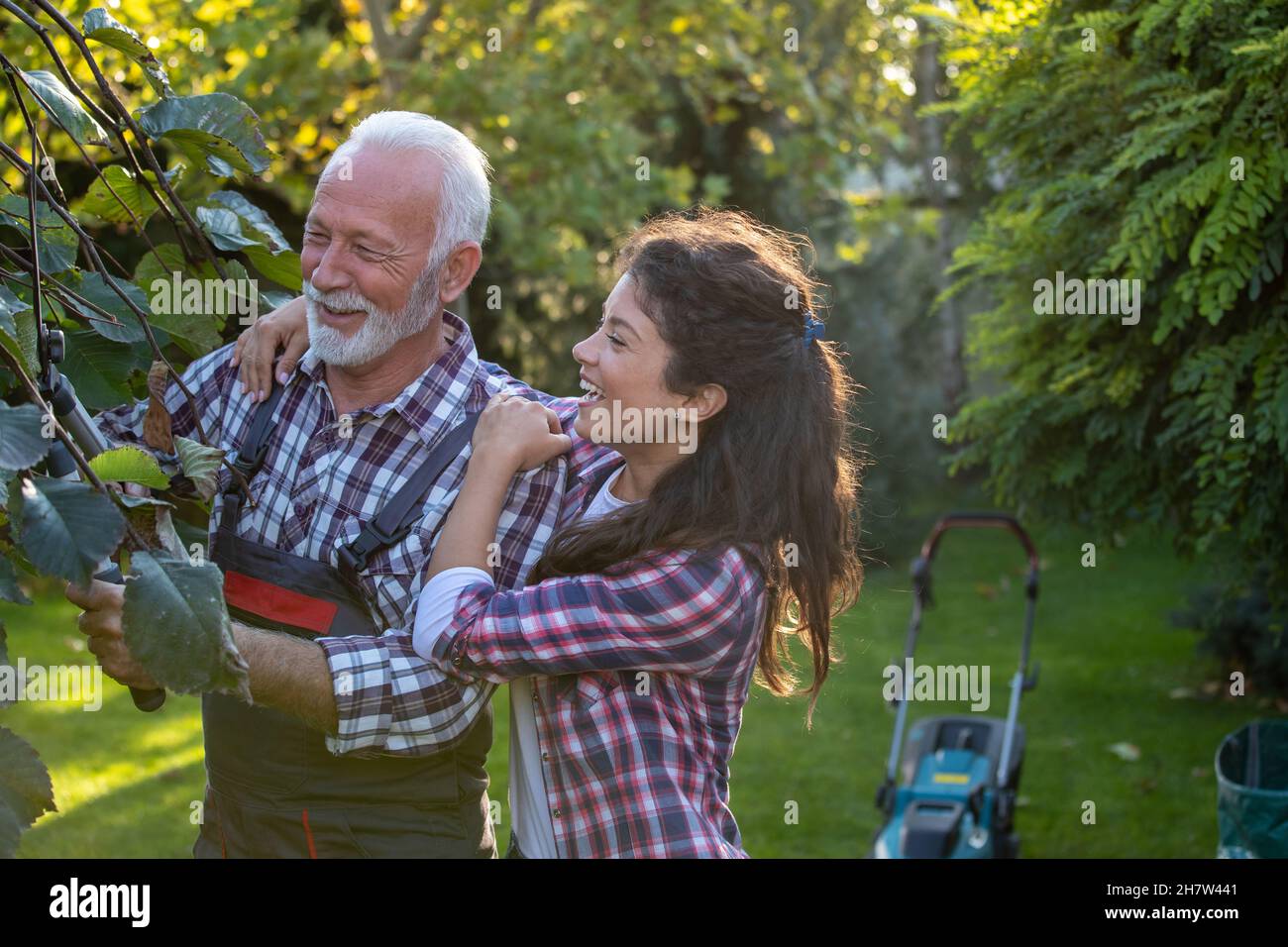 Uomo anziano che pota rami di albero mentre figlia graziosa che lo abbraccia in giardino Foto Stock