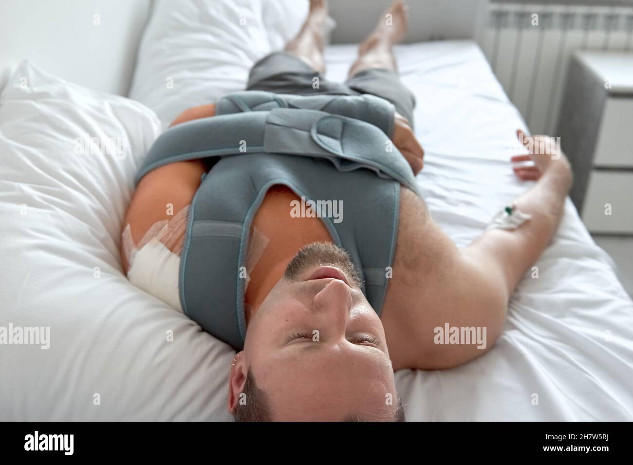 Un giovane paziente con catetere contagocce adagiato sul letto del reparto ospedaliero. Traumatologia intervento medico in caso di emergenza. Frattura della spalla del bendaggio elastico Foto Stock