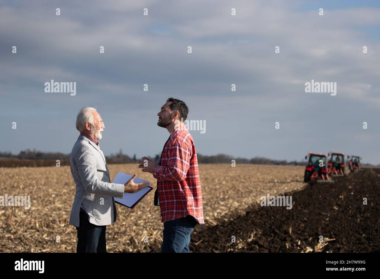 Uomo d'affari esperto che parla con l'agricoltore più giovane sul campo davanti ai trattori in autunno Foto Stock