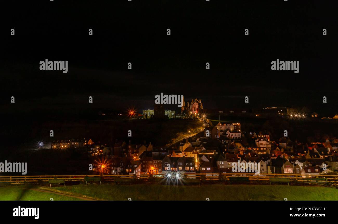 Whitby di notte. Le case fiaccano la collina con le finestre illuminate e una chiesa è illuminata sulla cima della scogliera. Un cielo notturno nuvoloso è sopra. Foto Stock