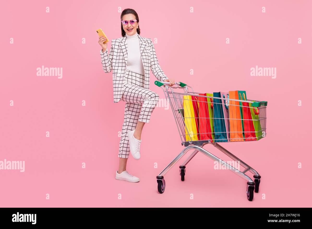 Immagine a grandezza naturale di donna sorridente funky in occhiali andare shopping chattare in smartphone isolato su sfondo rosa Foto Stock
