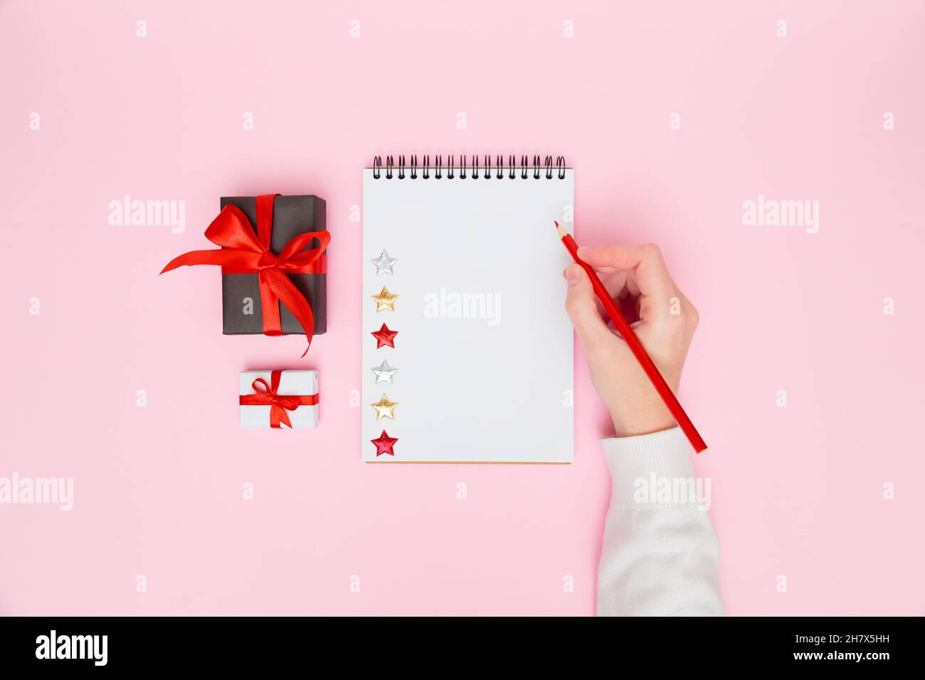 Sfondo accogliente Natale. Scrittura a mano femminile in taccuino bianco con scatole regalo con arco rosso su rosa pastello. Pianificazione del nuovo anno, obiettivi, elenco di cose da fare o Foto Stock