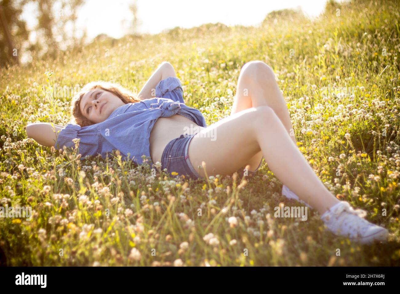 Giovane countrygirl bionda in denim con bottoni e calzini caldi jean in posa durante l'ora d'oro estiva all'aperto guardando seducente la fotocamera Foto Stock
