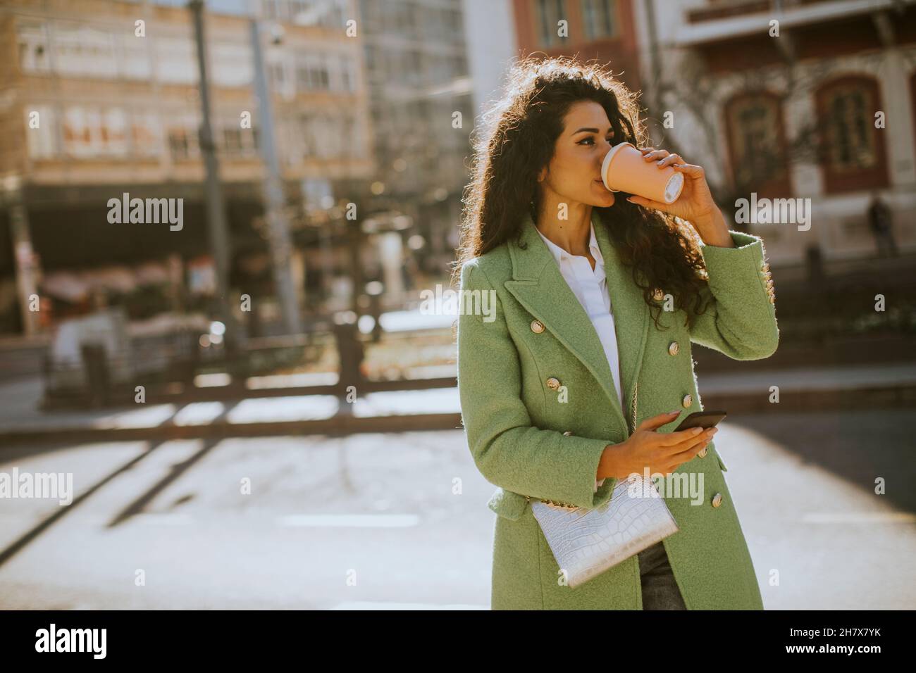 Donna piuttosto giovane che usa lo smartphone per strada e tiene il caffè da asporto Foto Stock