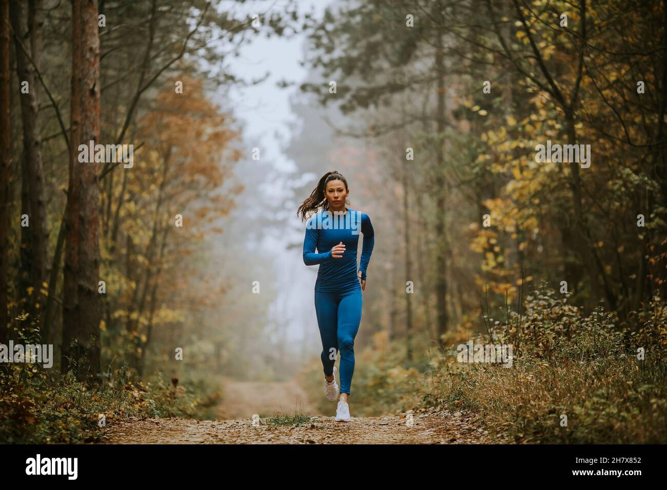 Giovane donna in tuta da pista blu che corre verso la macchina fotografica sul sentiero forestale in autunno Foto Stock