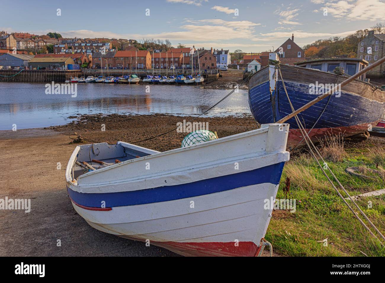 Un dinghy rosso, bianco e blu è ormeggiato su una slitta. Una vecchia barca dilapidata è dietro ed edifici e yacht sono sulla riva lontana. Foto Stock