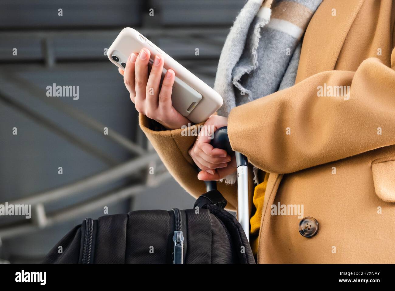 Il passeggero femminile in giacca con sciarpa effettua il check-in del volo dal telefono cellulare tenendo una grande valigia nel corridoio della vista vicina aeroporto internazionale Foto Stock