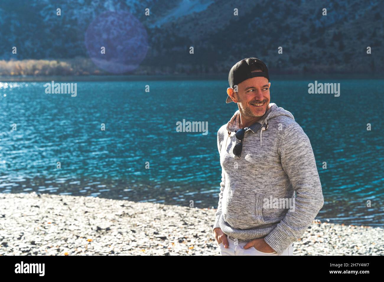 Uomo in piedi di fronte al lago di montagna con cappuccio a sfera sullo sguardo lontano Foto Stock