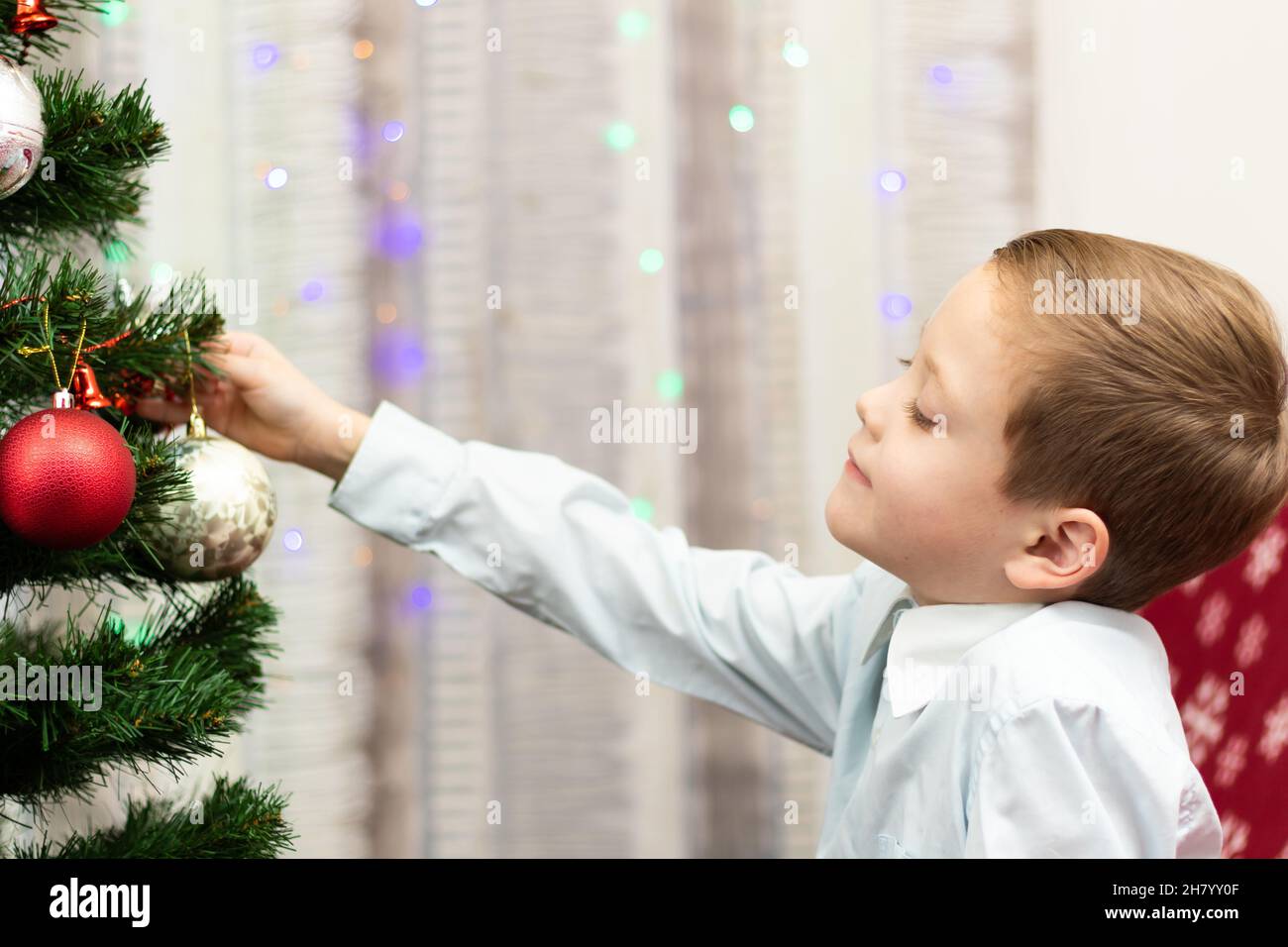 Un ragazzo di sette anni in una camicia blu decora un albero di Natale con giocattoli a casa la vigilia di Capodanno su uno sfondo di ghirlande. Messa a fuoco selettiva. Foto Stock