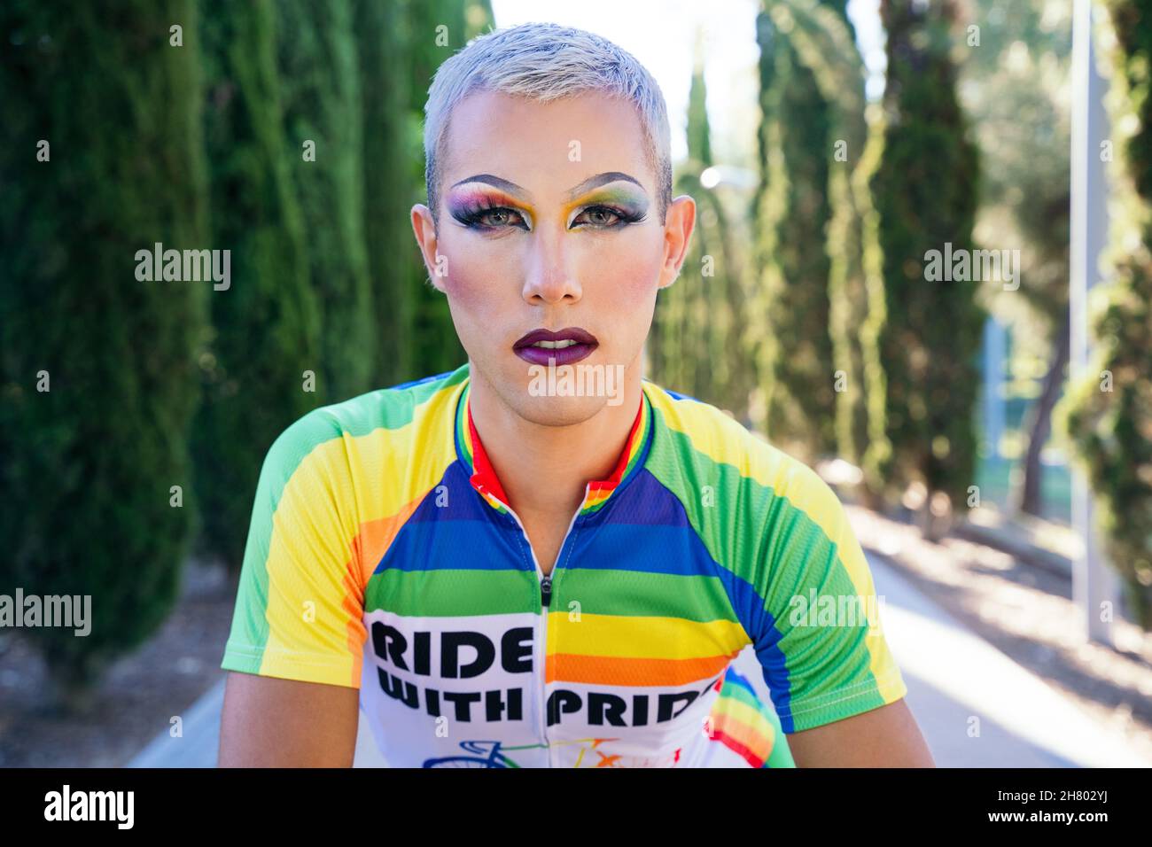 Ciclista gay seria con trucco luminoso in abbigliamento sportivo con  bandiera LGBT guardando la macchina fotografica mentre si siede in  bicicletta su strada asfaltata durante la maratona in ci Foto stock -