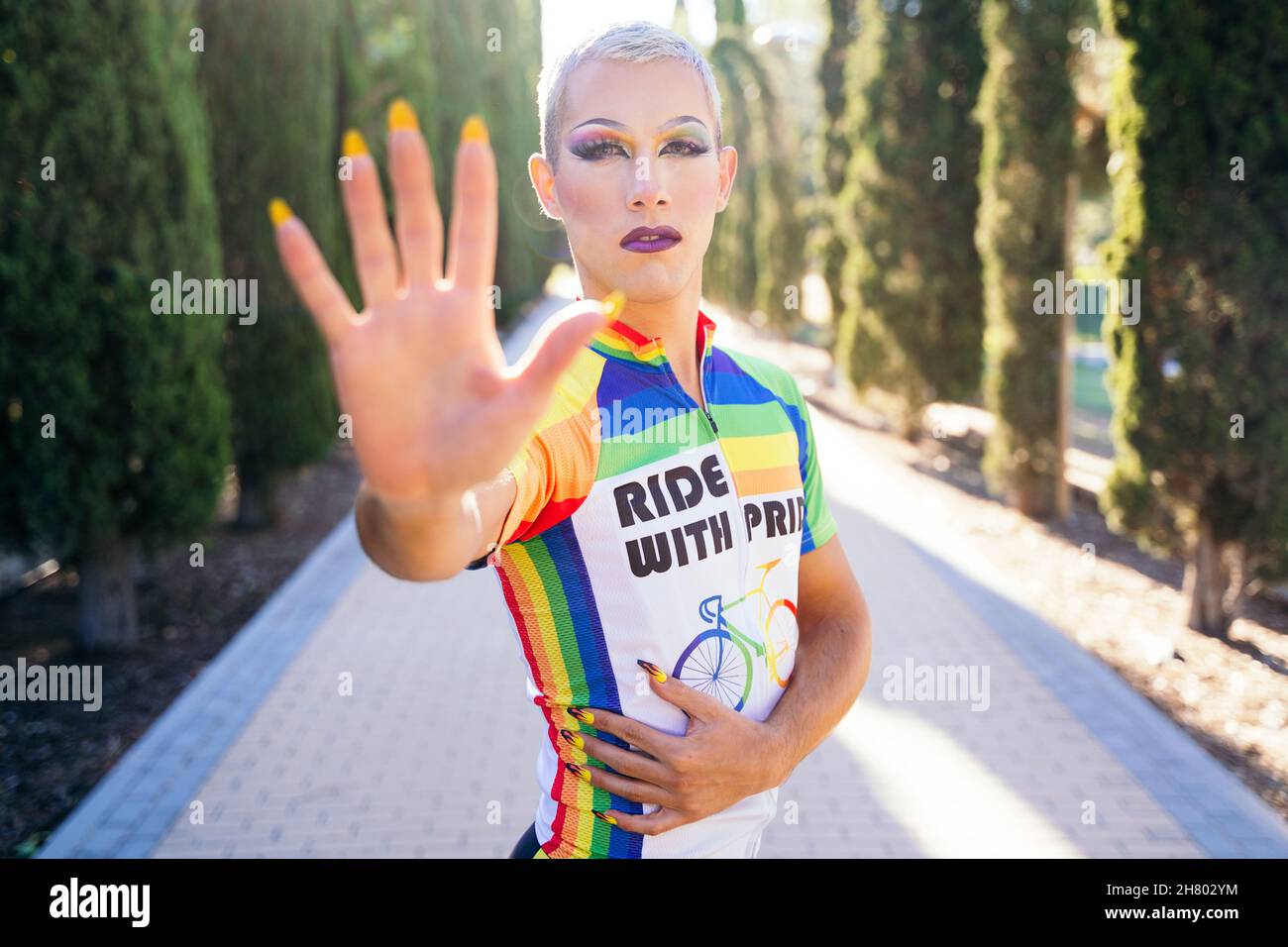 Uomo omosessuale in abbigliamento sportivo con arcobaleno LGBT che mostra  mano a macchina fotografica mentre si trova su una strada soleggiata  durante il giro in bicicletta Foto stock - Alamy