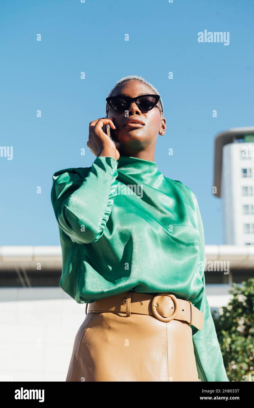 Fidata giovane afroamericana donna d'affari con capelli corti tinti in abiti alla moda e occhiali da sole che parlano sul telefono cellulare mentre si trova in piedi sulla strada Foto Stock