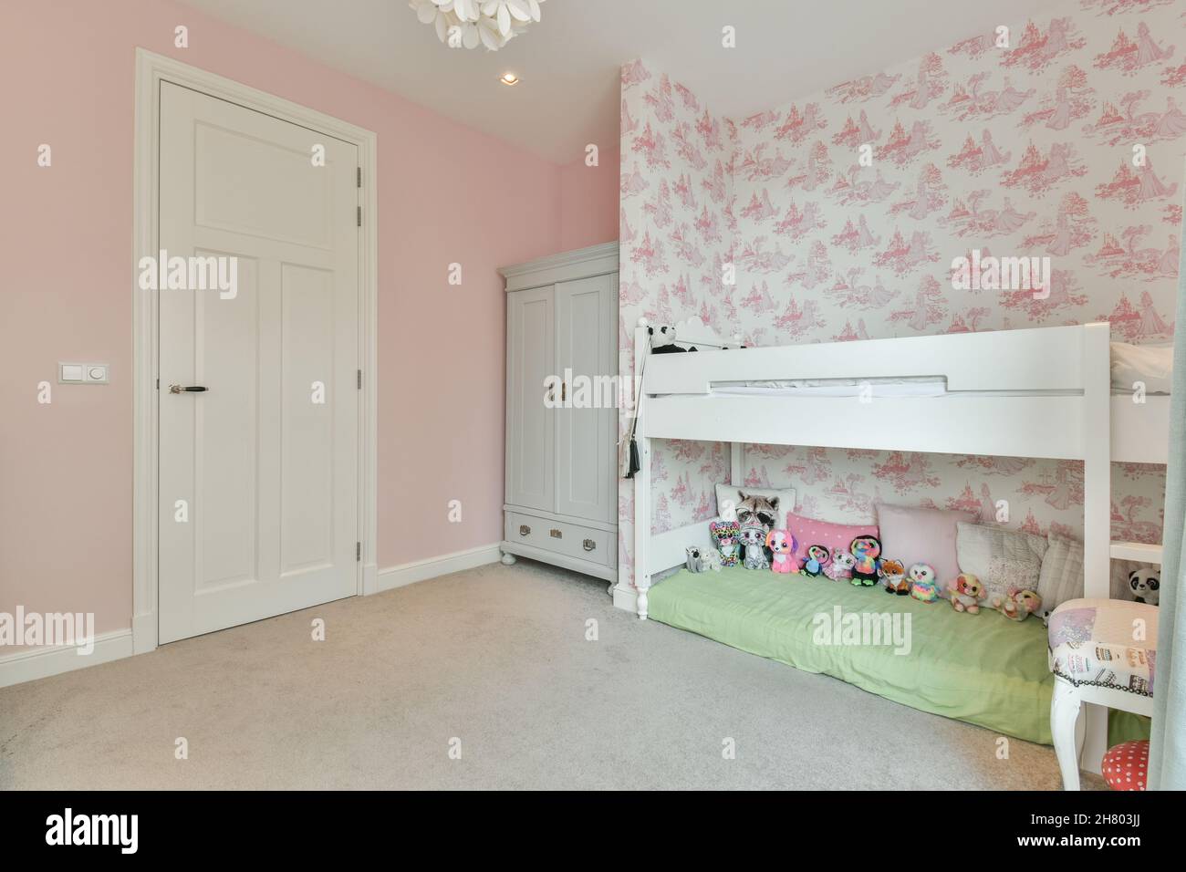 Interno di spaziosa camera da letto per ragazza con carta da parati rosa arredato con armadio in legno bianco vicino letto con diversi giocattoli e libreria Foto Stock