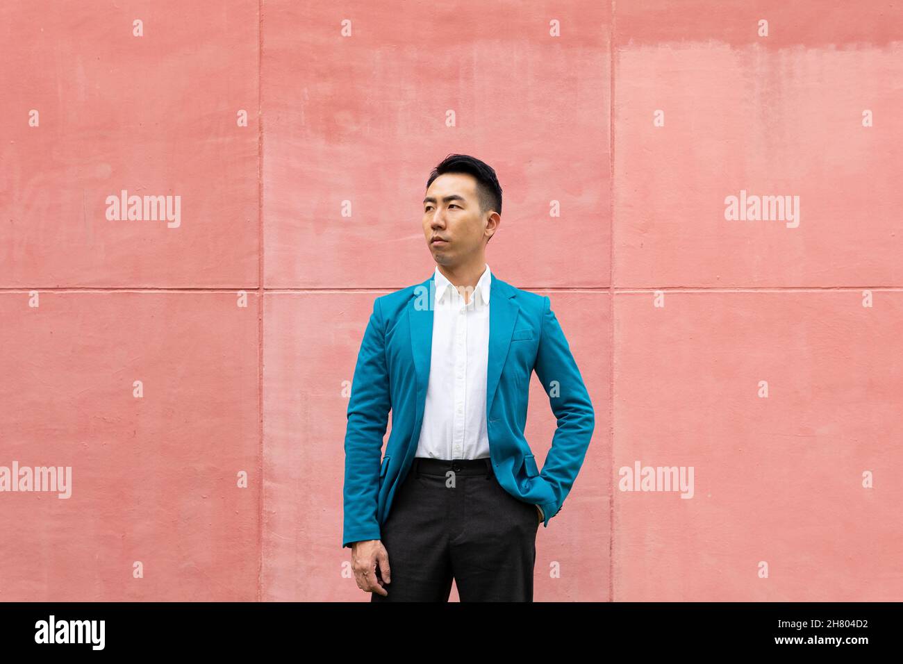 Uomo asiatico sicuro in abbigliamento formale in elegante giacca blu vestito guardando lontano mentre si trova in strada vicino alle mura luminose della città Foto Stock