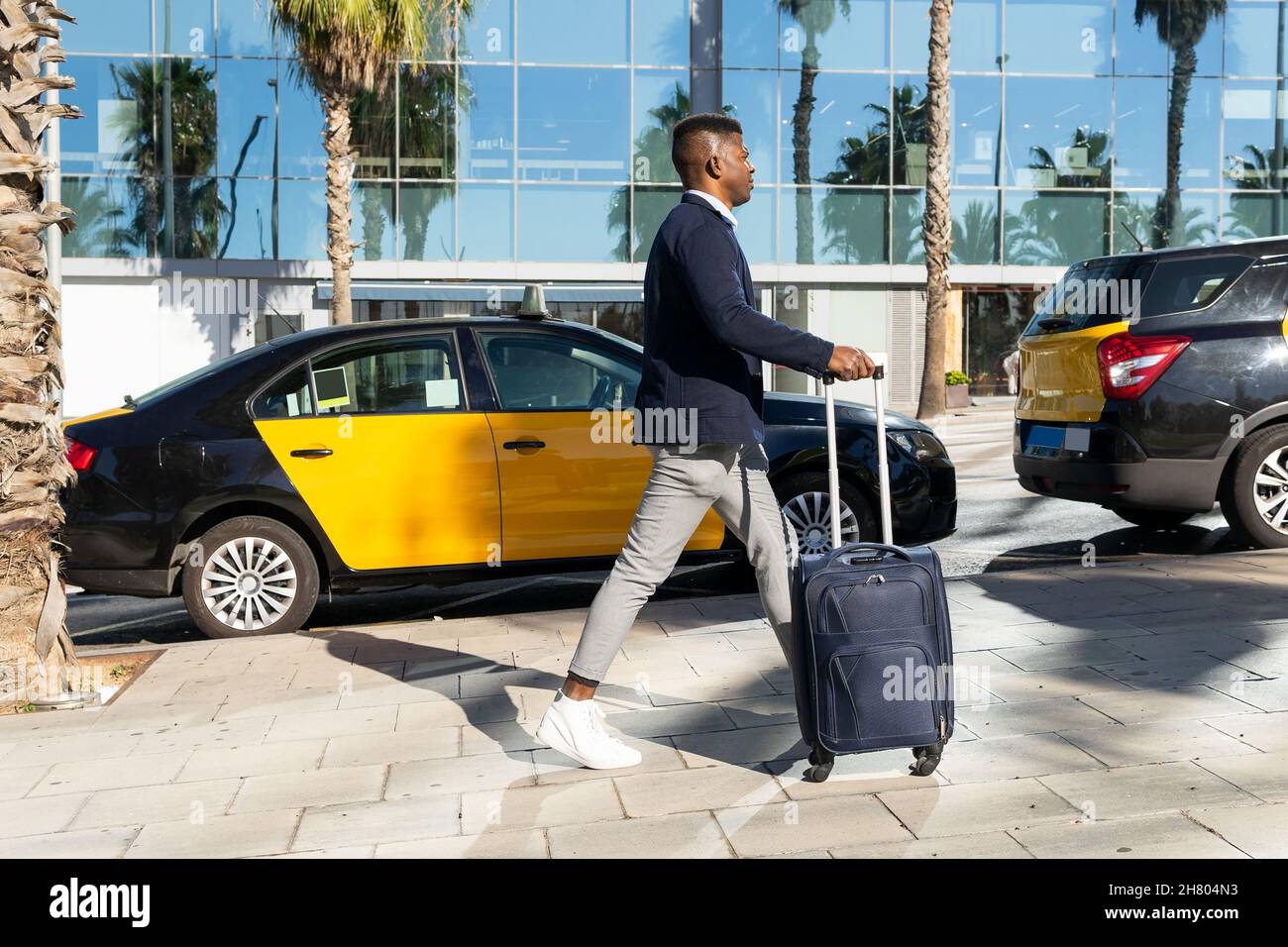 Vista laterale del corpo del turista maschile afroamericano in abbigliamento formale passeggiando con valigia sul marciapiede durante il viaggio di lavoro Foto Stock