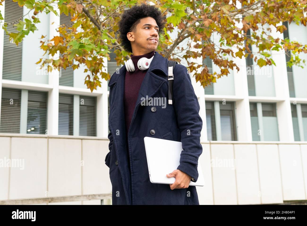 Fidato studente africano americano maschio con cuffie sul collo in piedi con netbook sotto l'albero autunnale e guardare via Foto Stock