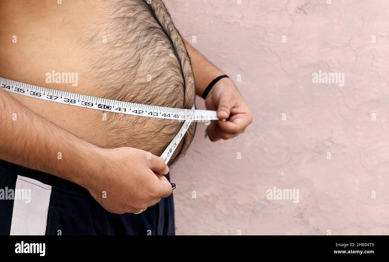 Un uomo asiatico misura il suo ventre grasso con un nastro di misurazione su uno sfondo semplice Foto Stock