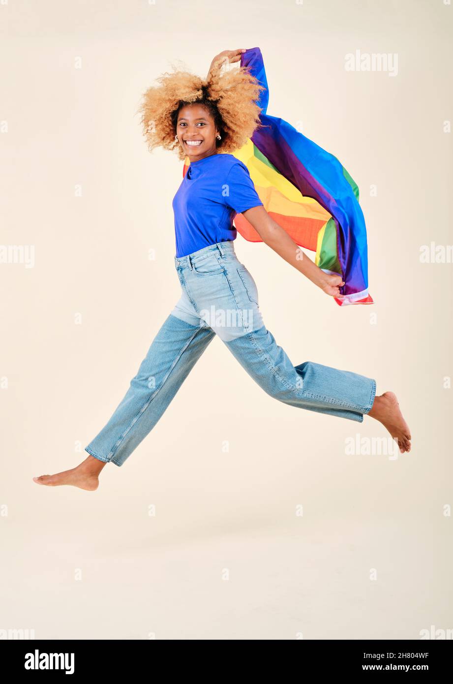 Felice giovane afro donna saltando con un arcobaleno gay orgoglio bandiera su uno sfondo isolato. Foto Stock