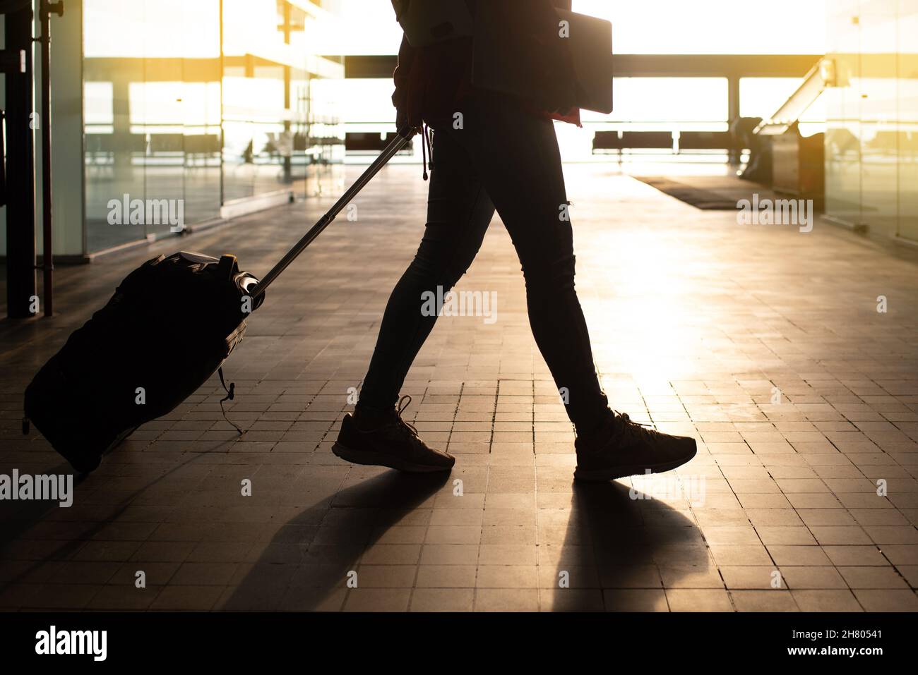 Ritagliate un viaggiatore irriconoscibile con un computer portatile in mano e una valigia che passeggiando nel moderno terminal pubblico dell'aeroporto con luce solare intensa prima del volo Foto Stock
