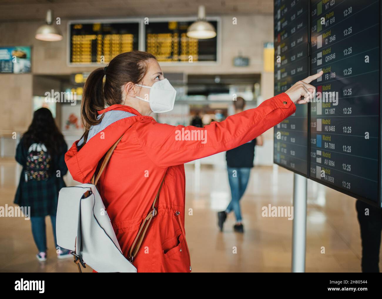 Vista laterale del viaggiatore femminile in maschera protettiva in piedi vicino al cartello con orario dei voli nel terminal dell'aeroporto prima della partenza Foto Stock