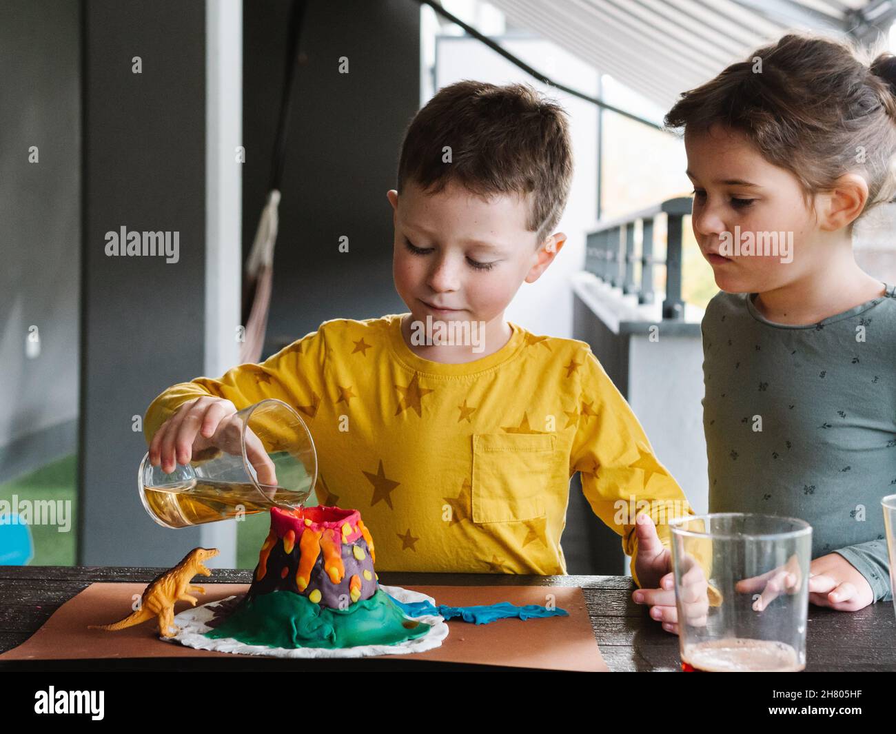 Adorabile ragazzo focalizzato versando liquido rosso nel cratere del vulcano creato con plastilina colorata con la sua sorellina Foto Stock