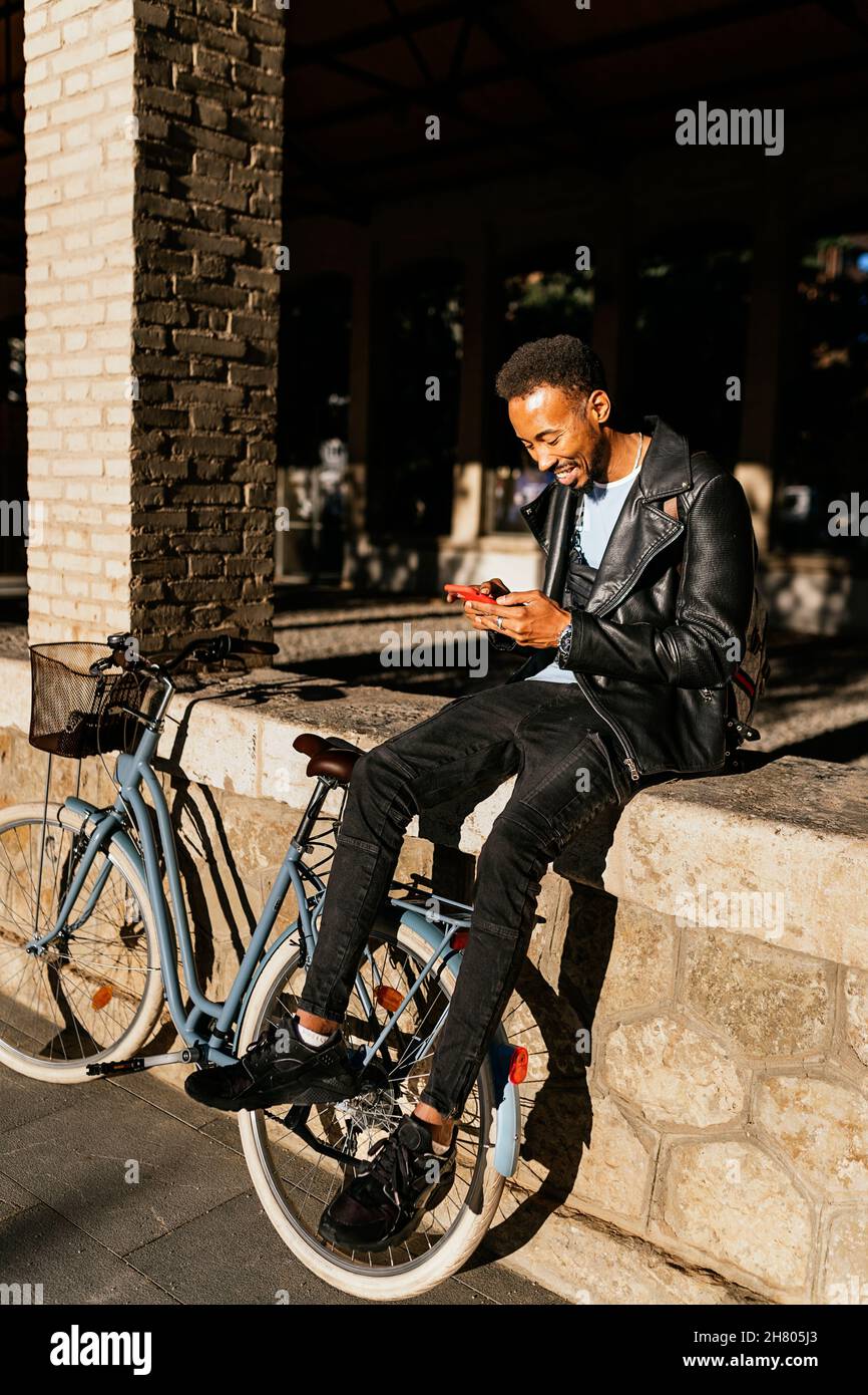 Vista laterale del corpo del allegro maschio afroamericano con zaino seduto con bicicletta sulla strada lastricata vicino all'edificio in città Foto Stock