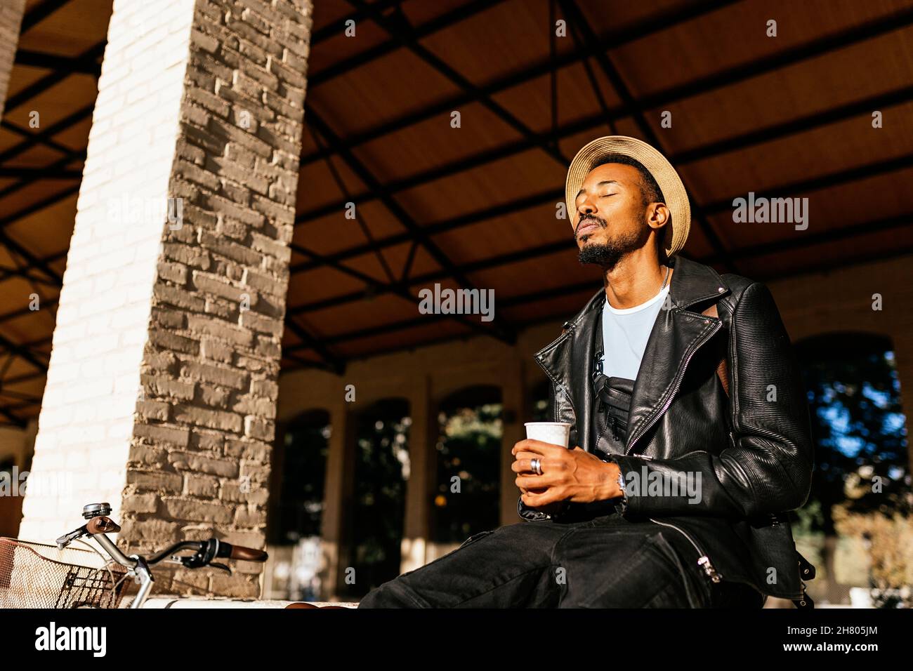 Calmo maschio afroamericano in cappello con bevanda da asporto seduta sulla terrazza dell'edificio con gli occhi chiusi mentre si gode la giornata di sole Foto Stock