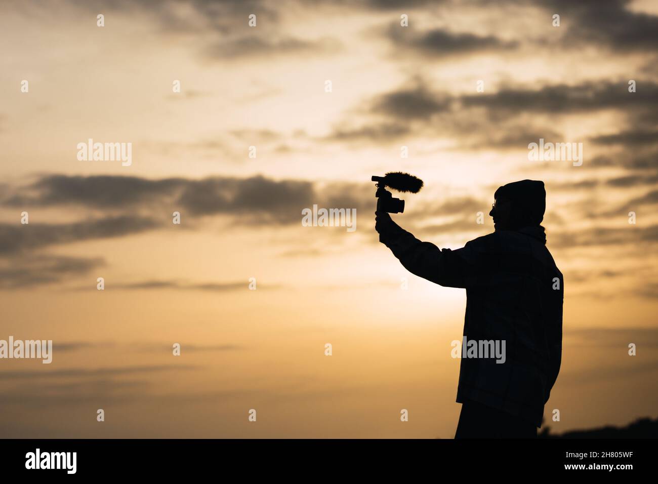 Vista laterale silhouette di giovane blogger femminile irriconoscibile in video di scatto cappello su fotocamera digitale contro il cielo del tramonto in campagna Foto Stock