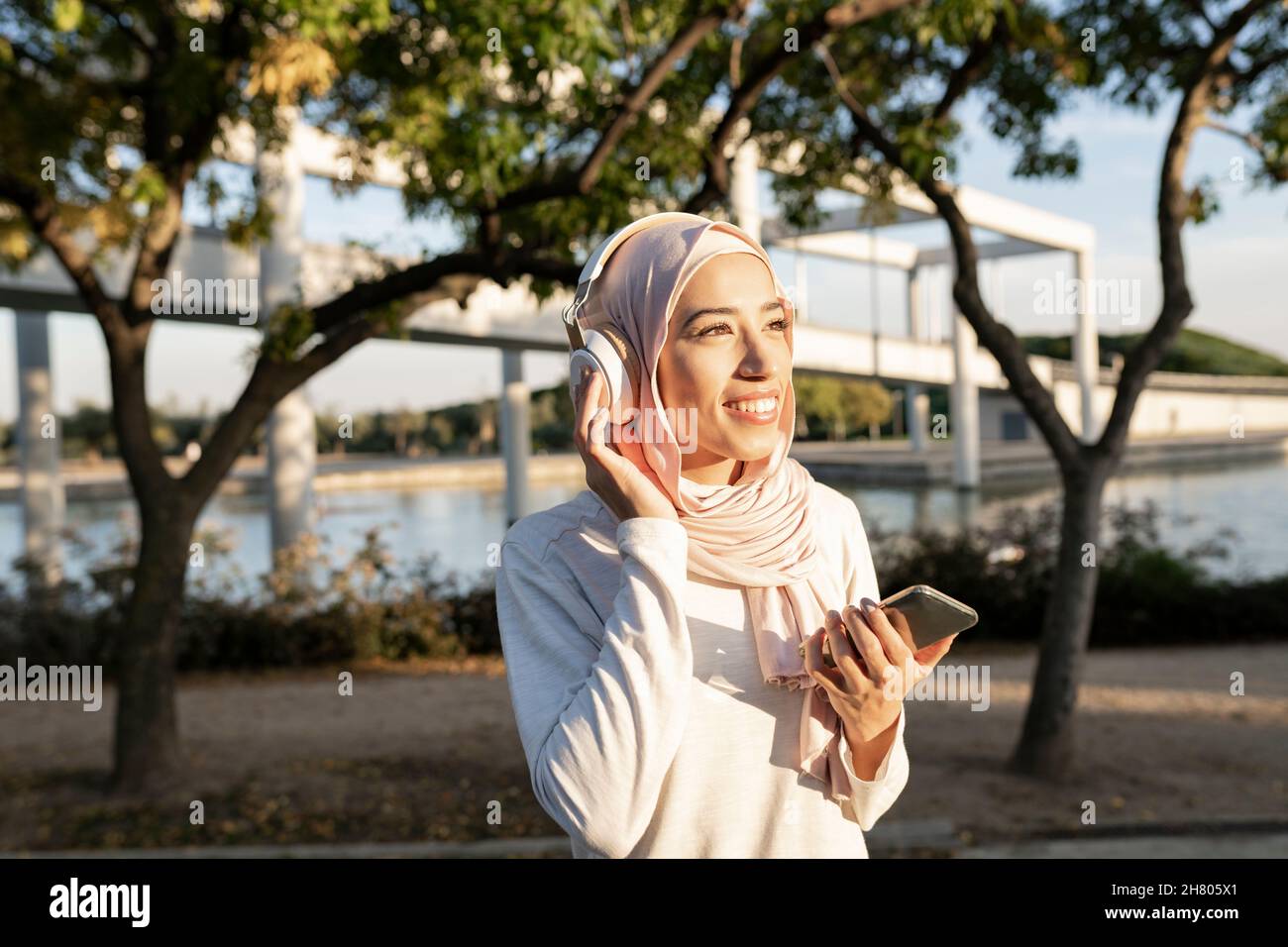 Positiva femmina araba in hijab seduta su scala di pietra vicino zaino e utilizzando il cellulare dopo l'allenamento all'aperto e musica listenig su heph Foto Stock