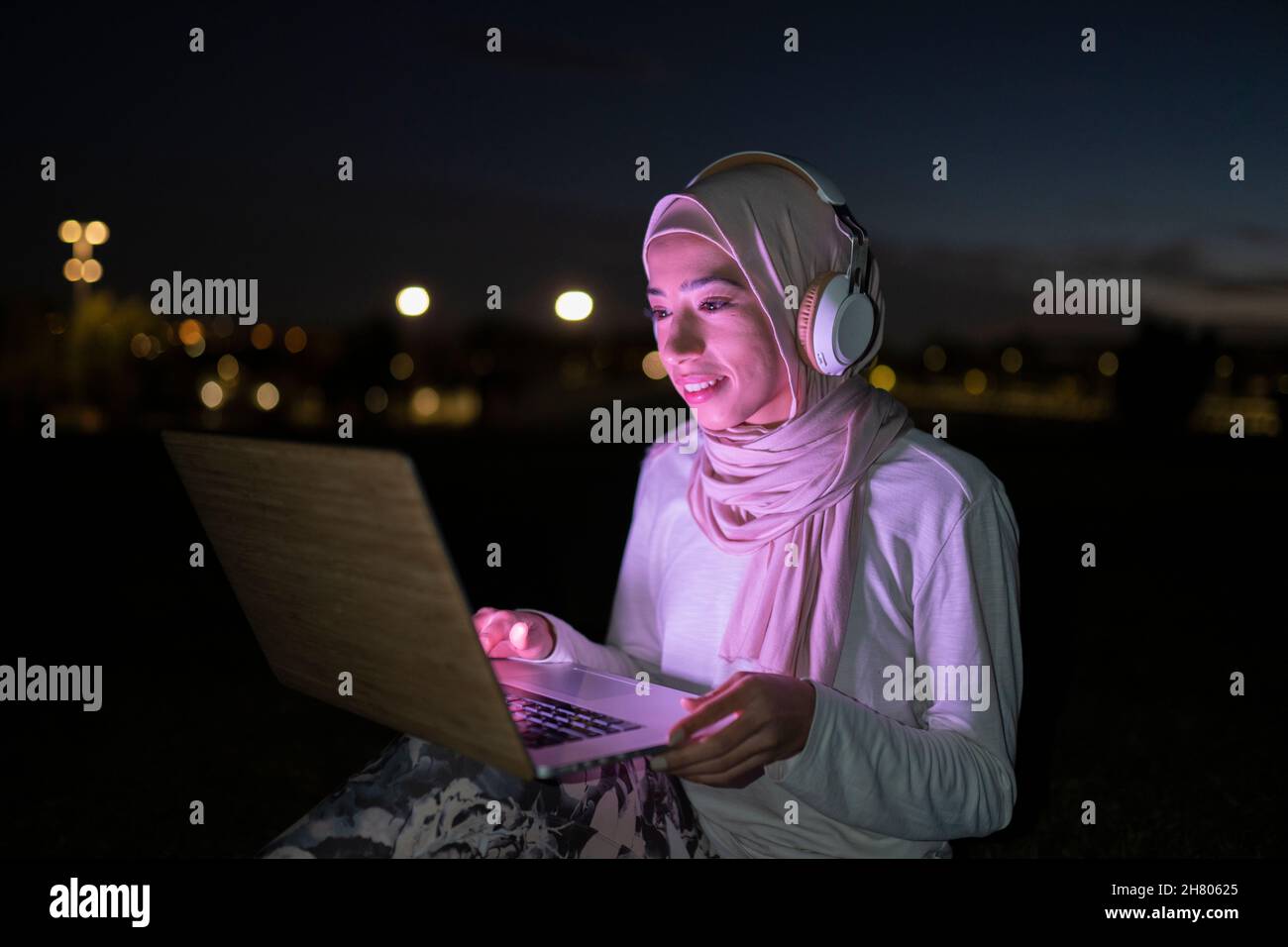 Positiva femmina araba in hijab utilizzando cuffie wireless e guardare video su netbook con luce viola nel buio Foto Stock