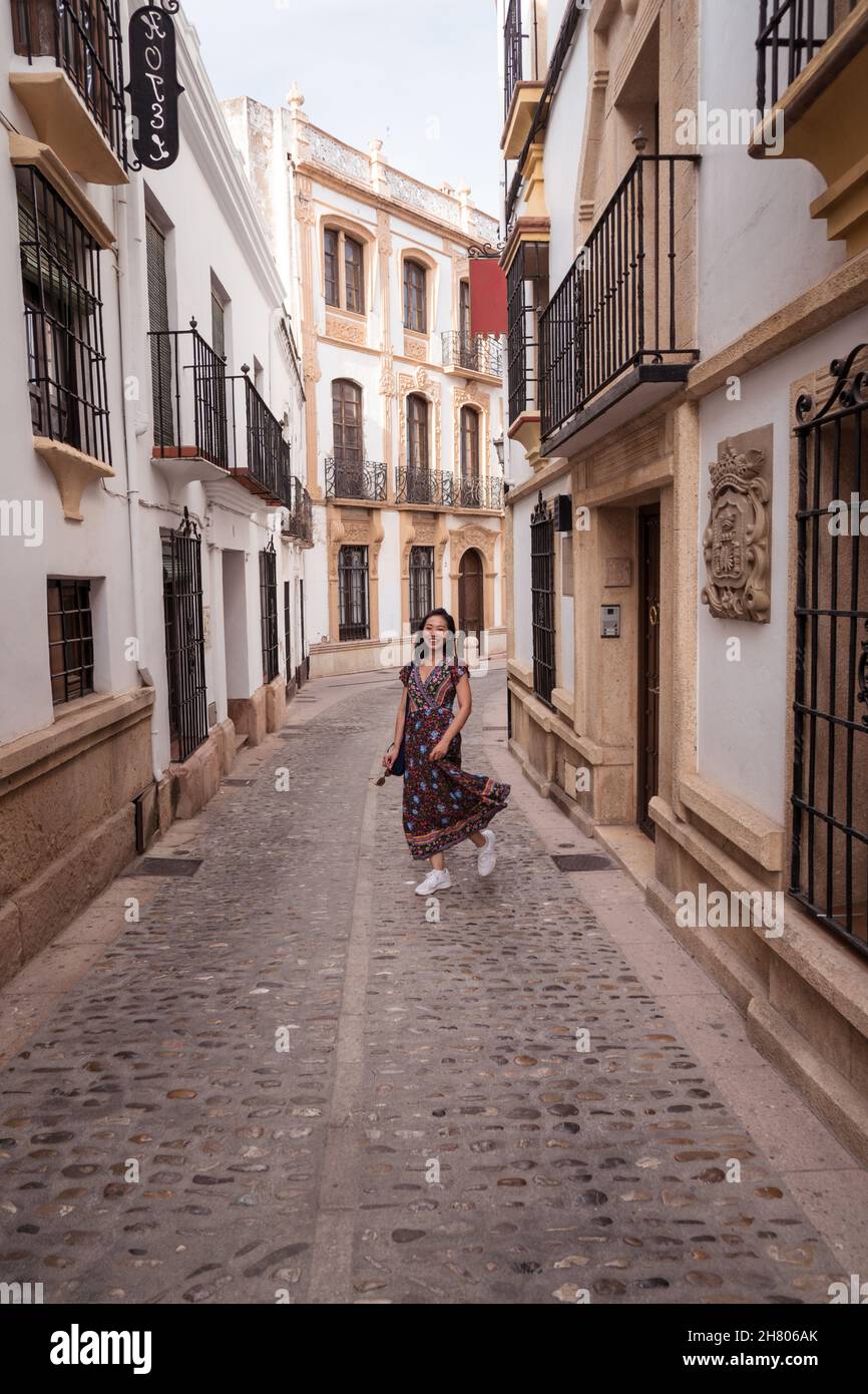 Pieno corpo di allegro viaggiatore asiatico femminile in abito guardando la macchina fotografica mentre si trova in piedi su strada lastricata vicino vecchie case Foto Stock