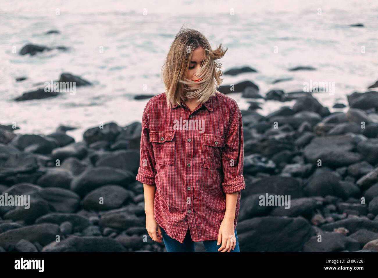 Il viaggiatore femminile è calmo e premuroso, con capelli volanti in piedi sulla costa sassosa e occhi chiusi vicino al mare increspato nella zona costiera durante il viaggio nella natura Foto Stock