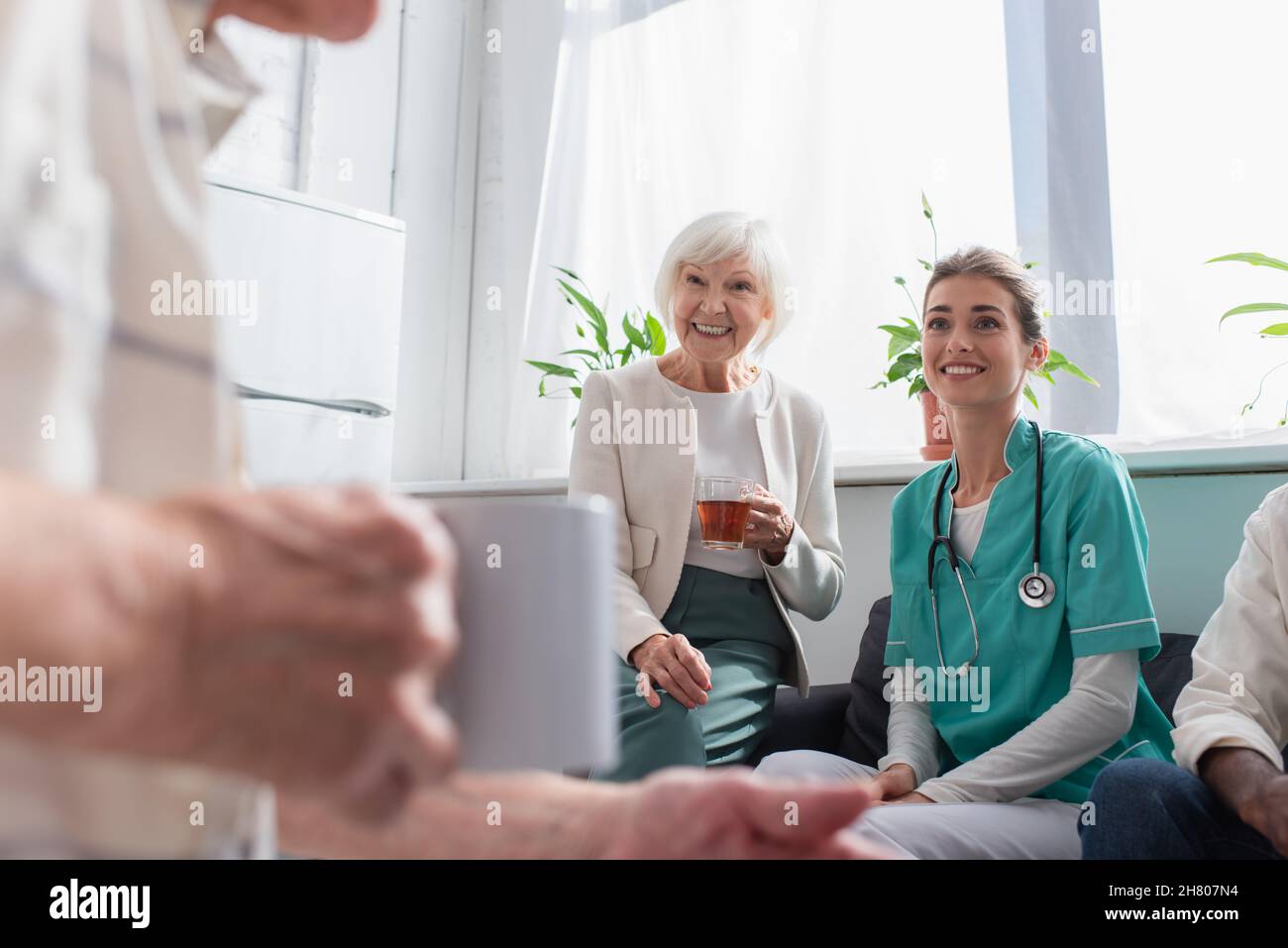 Donna positiva con tè e infermiera guardando l'uomo anziano in casa di cura Foto Stock