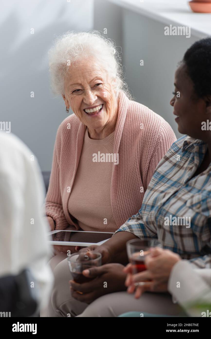 Sorridente paziente anziano che tiene un tablet digitale vicino ad amici interrazziali con tè in casa di cura Foto Stock