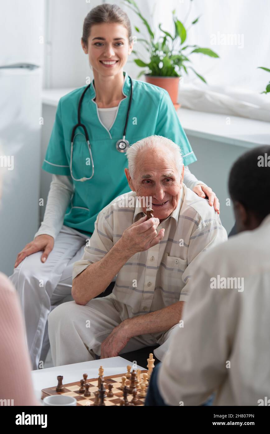 Uomo anziano che tiene la figura di scacchi vicino infermiera sorridente e amico afroamericano in casa di cura Foto Stock
