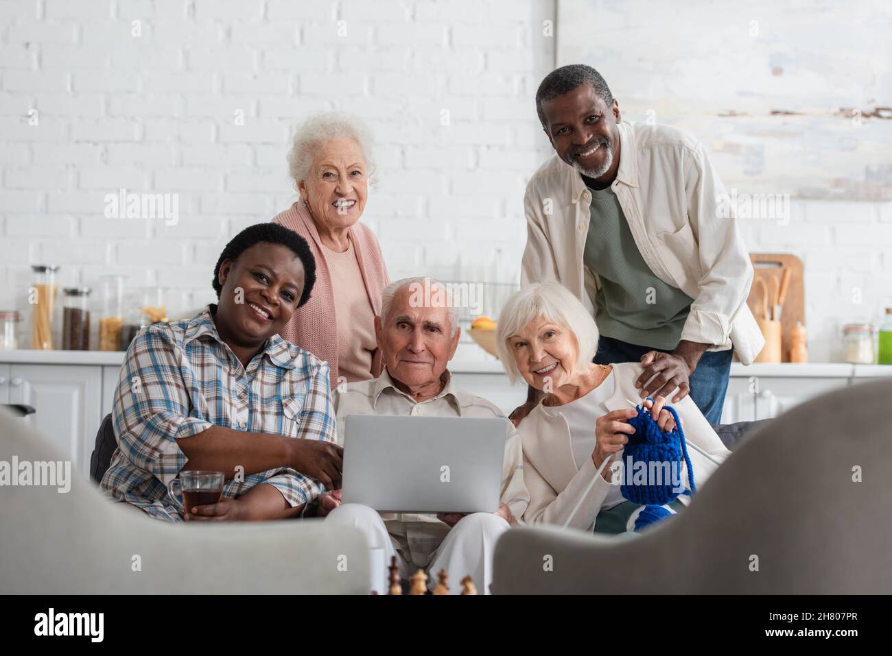 Pensionati multietnici positivi con filo, tè e computer portatile guardando la macchina fotografica in casa di cura Foto Stock