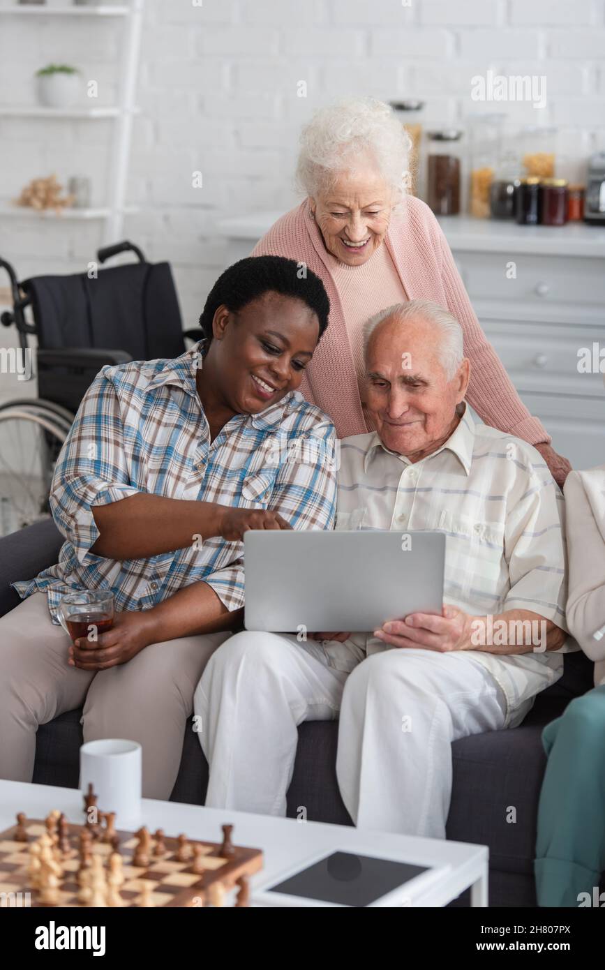 Donna afroamericana che tiene il tè vicino ad amici anziani con computer portatile e scacchi in casa di cura Foto Stock
