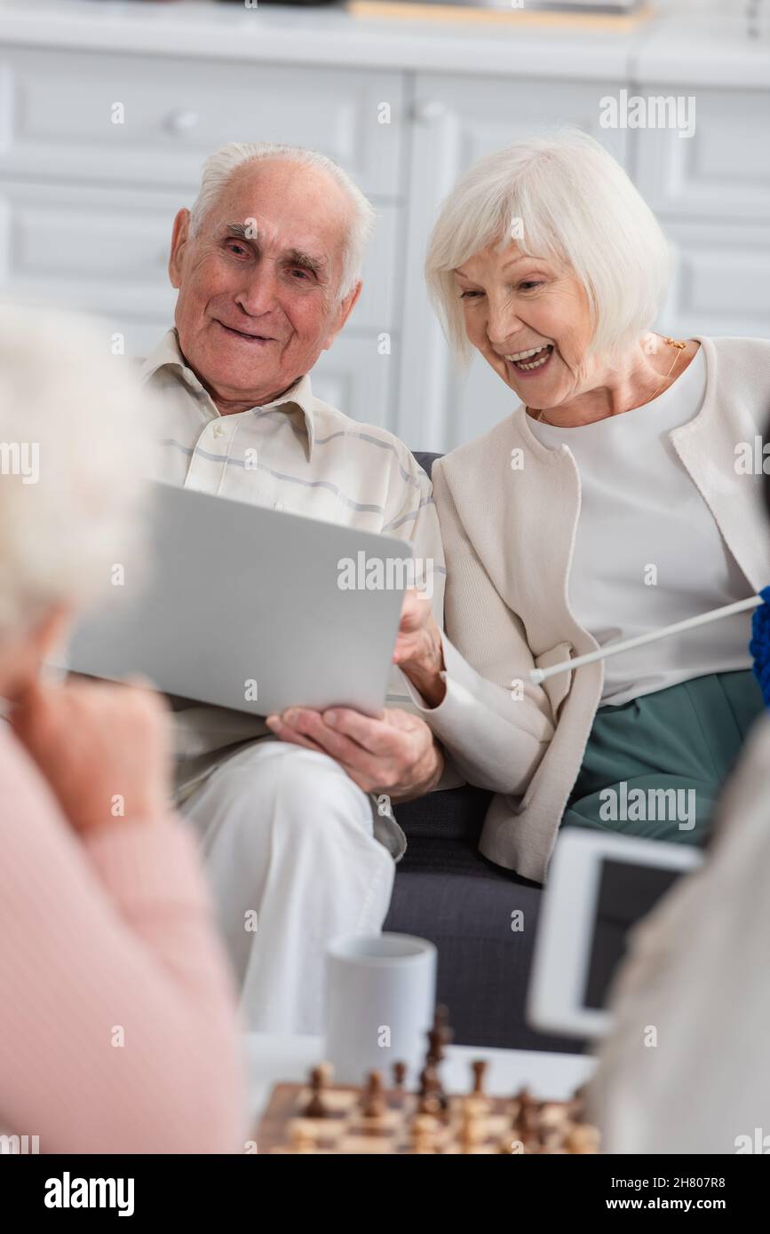 Donna anziana positiva con filo che guarda il laptop vicino ad un amico e scacchi sfocati in casa di cura Foto Stock