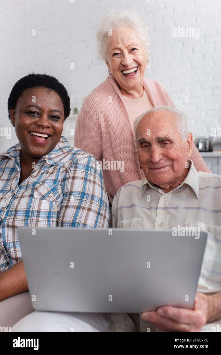Amici multietnici positivi con un computer portatile che guarda la macchina fotografica in casa di cura Foto Stock