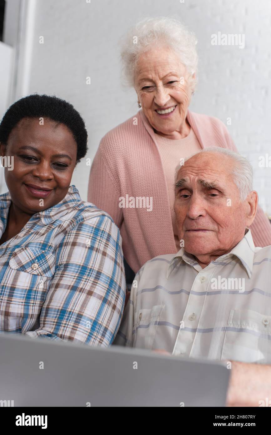Anziani pensionati multietnici che guardano un notebook offuscato in casa di cura Foto Stock